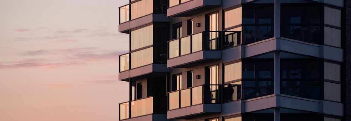 A modern apartment building with glass balconies reflecting a soft pink sky